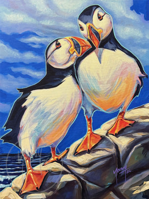 Lovebirds by Adam Young, two Atlantic puffins leaning together on grey rocks against a bright blue sky, with surf below